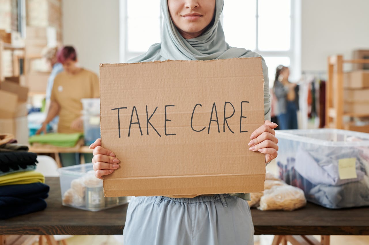 Smiling volunteer in hijab holding a 'Take Care' sign at a charity event indoors.